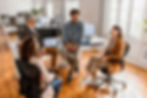 A casual meeting in an office with four colleagues laughing and talking in a relaxed atmosphere.