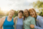Four older women smiling and embracing outdoors, enjoying a moment of friendship and happiness in a natural setting.