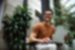 Smiling man sitting outdoors, holding a laptop, surrounded by lush green plants in a modern workspace.