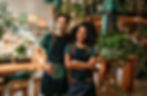 Two plant shop owners wearing aprons standing confidently amidst lush greenery and wooden furniture.