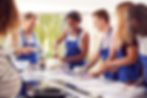 A group of people, wearing blue aprons, smiling and working together during a cooking class.