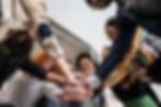 A diverse team joins hands in a show of unity and teamwork, smiling and looking up at each other.