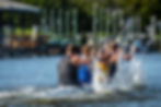 Four kayakers vigorously paddling in a race, splashing water around in a sunny setting.