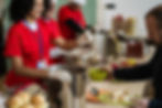 Volunteers in red shirts serving meals to people in a community kitchen