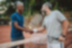 Two men shaking hands at the net after a tennis match, holding their rackets and smiling.