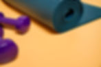 Close-up of a rolled yoga mat and two purple dumbbells on an orange surface.