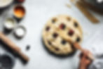 Hands applying egg wash to a homemade fruit pie with ingredients scattered on a marble countertop.