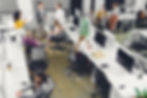 A group of people in a collaborative office space discussing work, surrounded by computer stations.
