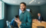 A confident man in a green shirt smiles while holding a smartphone in a modern conference room.
