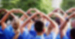 A crowd of people in blue shirts forming heart shapes with their hands in a green park