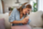 Joyful woman wearing headphones, laughing while looking at her smartphone on a couch.