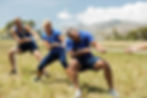 Three people in blue shirts pulling a rope in a competitive outdoor tug-of-war event.