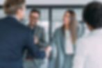 Business professionals shaking hands and smiling in a modern office, while colleagues watch and applaud in the background.