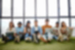 A diverse group of young adults sitting on artificial grass indoors, using laptops, tablets, and books for study or work.