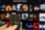 Close-up of a hand holding a TV remote with a blurred streaming service screen in the background.