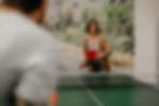 A woman joyfully playing ping pong, holding a red paddle, while another person watches.
