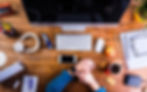 Overhead view of a rustic desk with a computer, gadgets, snacks, and a person checking their smartwatch.