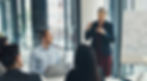 Businesswoman giving a presentation to a group in a conference room with a whiteboard.