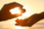 Two hands passing a heart-shaped object in front of a sunset on a beach, symbolizing love.