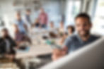 A man writing on a whiteboard, smiling, with a team of coworkers behind sitting on a table