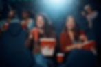 Two women laughing and holding popcorn while watching a movie in a theater.
