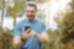 Middle-aged man smiling while using his smartphone in a sunny forest setting.
