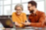 Two colleagues looking at a laptop while working and smiling, one of them an older woman, and the other a younger man.