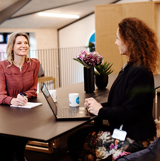 A smiling woman in a rose-colored blouse sitting at a desk with a notepad in front of a laptop, engaging in a conversation with another woman standing across, in a modern office environment with a "GoGift" promotional banner in the background.