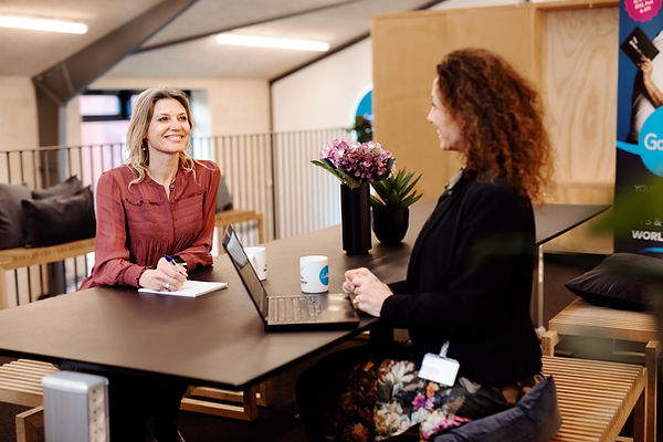A smiling woman in a rose-colored blouse sitting at a desk with a notepad in front of a laptop, engaging in a conversation with another woman standing across, in a modern office environment with a "GoGift" promotional banner in the background.